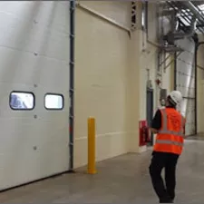 Worker in an orange safety vest and hard hat inspecting or operating a large industrial garage door inside a facility. 